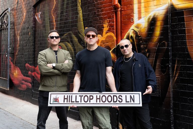 Australian hip-hop group Hilltop Hoods wearing sunglasses and standing in front of a painted brick wall holding a Hilltop Hoods Ln street sign.